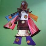 A paper collage figure with a mask-like face, wearing a colourful outfit with red, orange, and blue sleeves, purple trousers, and circular patterns, set against a green background.