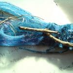 A bundle of blue fibres, twine, and sticks arranged on a white surface with faint handwritten text in the background.