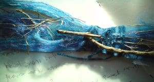 A bundle of blue fibres, twine, and sticks arranged on a white surface with faint handwritten text in the background.