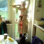 A person wearing an apron stands by a glass door, holding up a hanging decoration made of leaves, flowers, and branches. Another person sits at a nearby table with a mug and crafting materials.