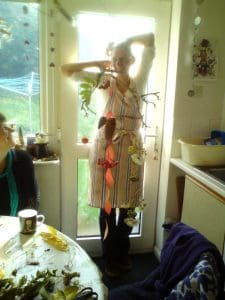 A person wearing an apron stands by a glass door, holding up a hanging decoration made of leaves, flowers, and branches. Another person sits at a nearby table with a mug and crafting materials.