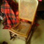 A wooden chair with a woven cane seat and backrest, partially damaged, sits on a carpeted floor next to a red checked blanket and a dark wooden cupboard.