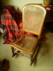 A wooden chair with a woven cane seat and backrest, partially damaged, sits on a carpeted floor next to a red checked blanket and a dark wooden cupboard.