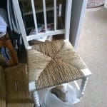 A white wooden chair with a woven seat is positioned near a plastic bag and stacked Amazon boxes, beside a small wooden table and some papers on the floor.