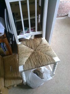 A white wooden chair with a woven seat is positioned near a plastic bag and stacked Amazon boxes, beside a small wooden table and some papers on the floor.