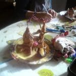 A person paints clay sculptures on a plate at a table covered with art supplies, fabric offcuts, and colourful materials.
