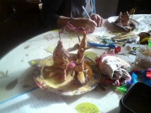 A person paints clay sculptures on a plate at a table covered with art supplies, fabric offcuts, and colourful materials.