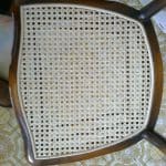 A wooden chair with a woven cane seat is shown from above, placed on a patterned floor and a chequered tablecloth.