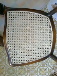 A wooden chair with a woven cane seat is shown from above, placed on a patterned floor and a chequered tablecloth.