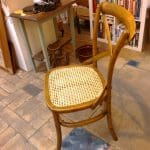 A wooden chair with a woven cane seat is positioned on a tiled floor near a small table and a bookcase.
