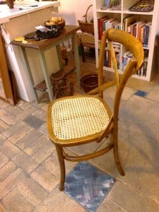 A wooden chair with a woven cane seat is positioned on a tiled floor near a small table and a bookcase.