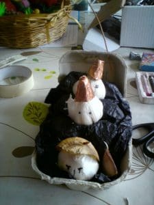 Three white onions with drawn faces and foil hats are placed in a carton lined with black paper, set on a table with craft supplies around.