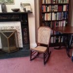A small wooden rocking chair sits on a pink carpet in front of a fireplace and next to a bookcase filled with books and a round wooden table.