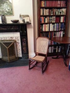 A small wooden rocking chair sits on a pink carpet in front of a fireplace and next to a bookcase filled with books and a round wooden table.