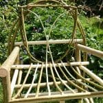 A handmade wooden garden chair with a decorative backrest and seat made from curved branches, placed on grass with greenery and flowers in the background.