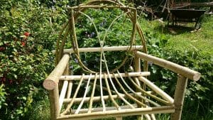A handmade wooden garden chair with a decorative backrest and seat made from curved branches, placed on grass with greenery and flowers in the background.