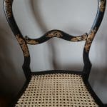 A black wooden chair with a decorative caned seat and gold floral detailing on the curved backrest and frame.