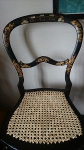A black wooden chair with a decorative caned seat and gold floral detailing on the curved backrest and frame.