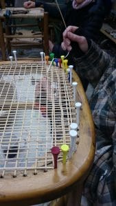 Close-up of a person weaving a cane chair seat, using colourful golf tees to secure the cane strips to the wooden frame.