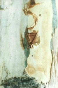 A close-up of a textured surface with peeling paint, exposing layers of green, beige, and brown, and intersecting lines resembling scratches or marks.