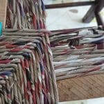 Close-up of a wooden chair frame being woven with multicoloured fabric strips, secured with a black and green clamp on one corner.