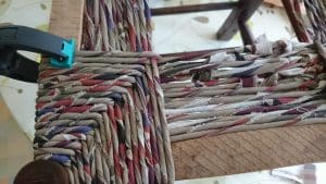 Close-up of a wooden chair frame being woven with multicoloured fabric strips, secured with a black and green clamp on one corner.