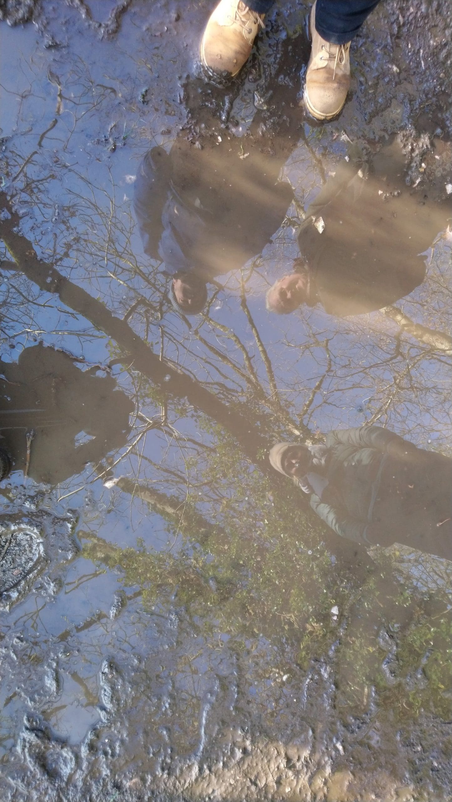 People’s reflections and surrounding trees are visible in a muddy puddle on a dirt path, with a pair of tan boots and dark shoes at the edge of the water.
