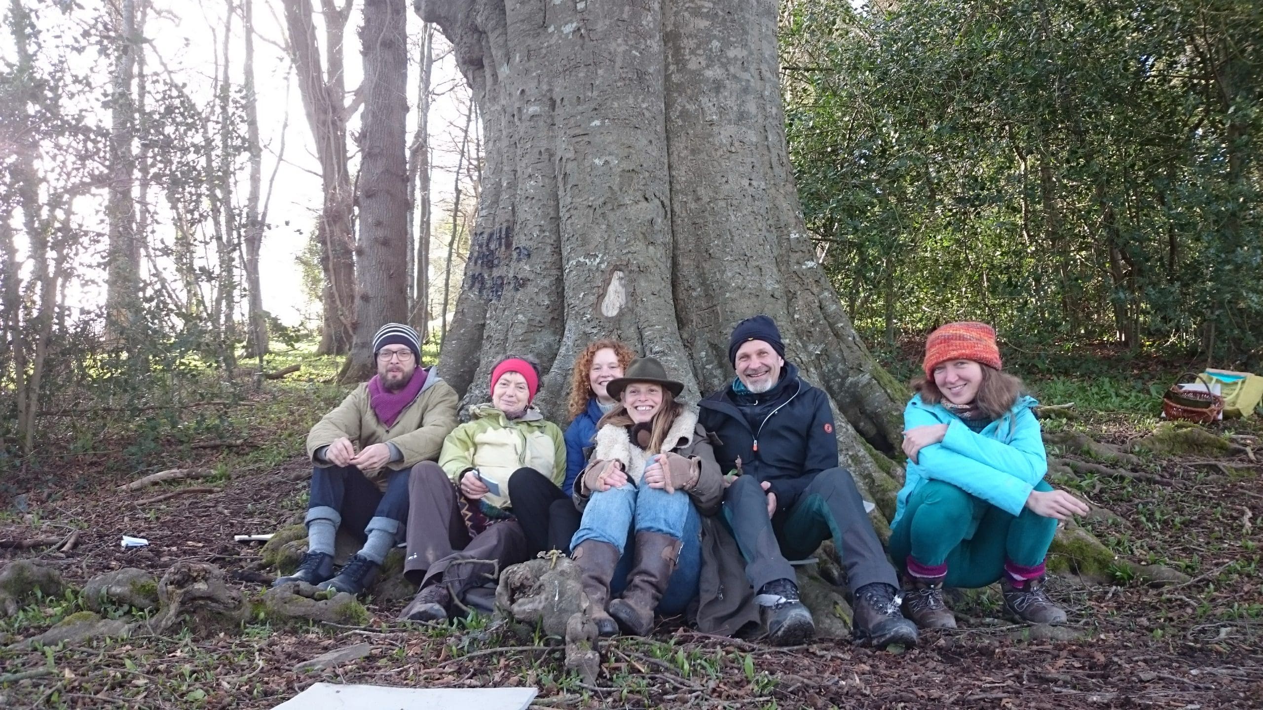 Six people sit side by side on the ground in front of a large tree trunk in a wooded area, all dressed in outdoor clothing and smiling.