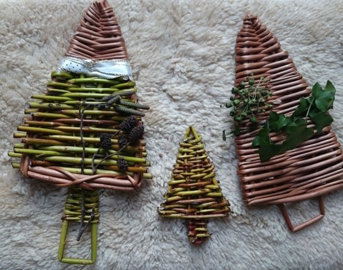 Three handmade wicker Christmas tree decorations, each decorated with natural materials, displayed on a light, textured fabric surface.