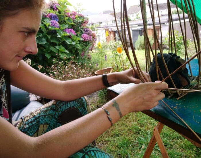 A woman weaves a basket with thin sticks at an outdoor table, surrounded by greenery and flowers.