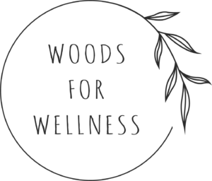 Black circular logo with the words "WOODS FOR WELLNESS" in uppercase letters and a branch with leaves extending from the circle's edge.