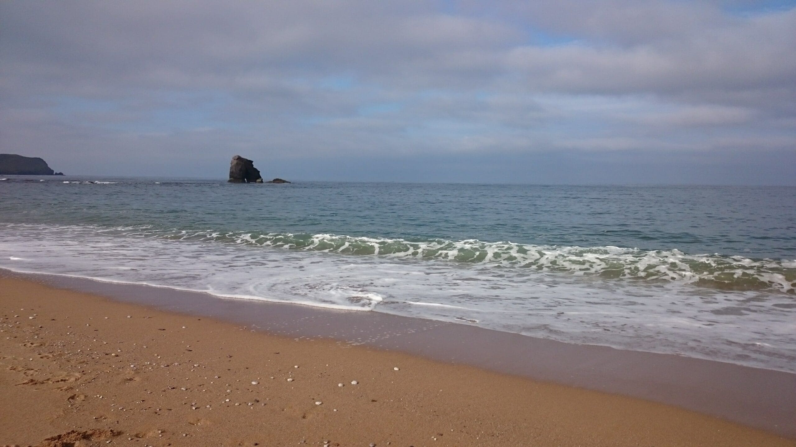 Sandy beach with gentle waves and a single large rock formation in the sea under a partly cloudy sky.