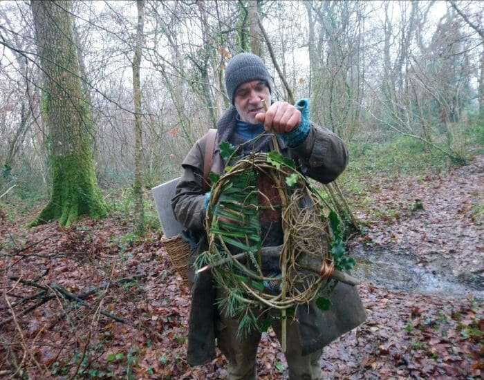 A person in a woolly hat and coat holds a handmade wreath of twigs and leaves in a damp, leaf-covered forest.