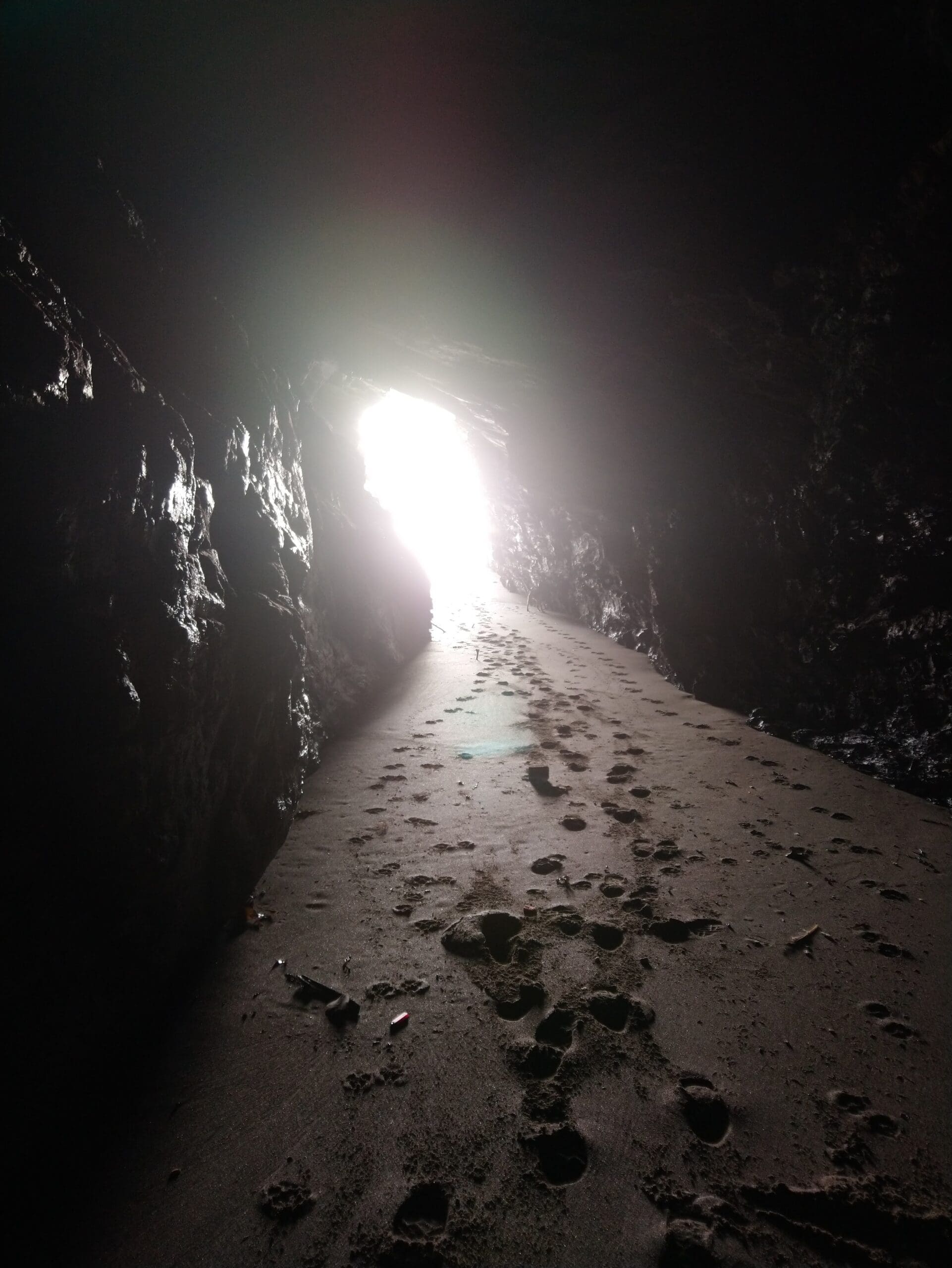 Footprints lead along sandy ground inside a dark cave towards a bright light at the cave’s entrance.