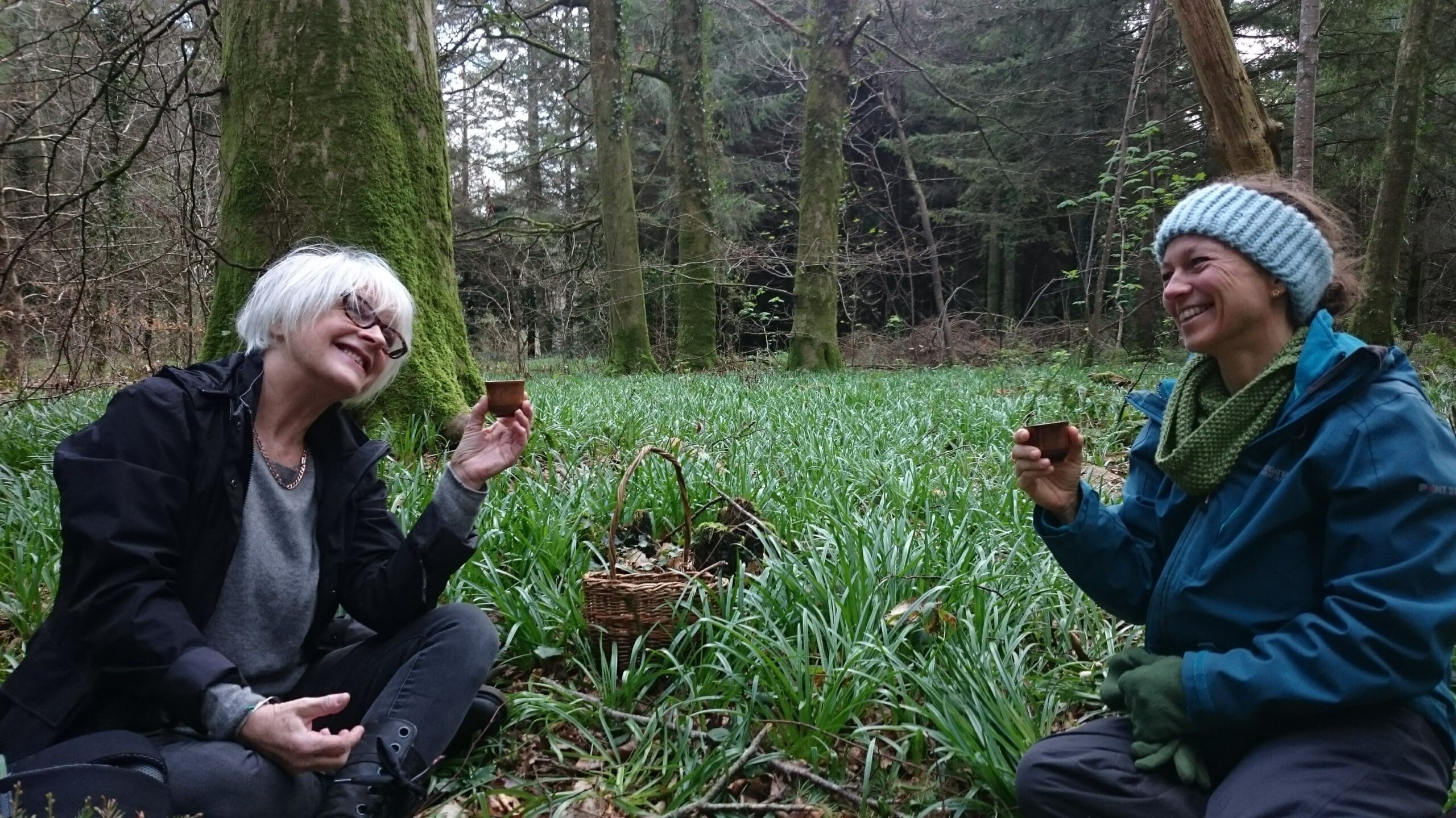 Two women sit on grass in a forest, smiling and holding small cups. A wicker basket sits between them. Both wear jackets; one has a headband and gloves, the other spectacles and white hair.