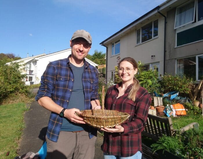 Two people standing outside near a bench, holding a woven basket together, with residential buildings and greenery in the background.