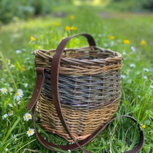 A woven basket with a leather strap sits on green grass surrounded by small white daisies, with a blurred natural background.