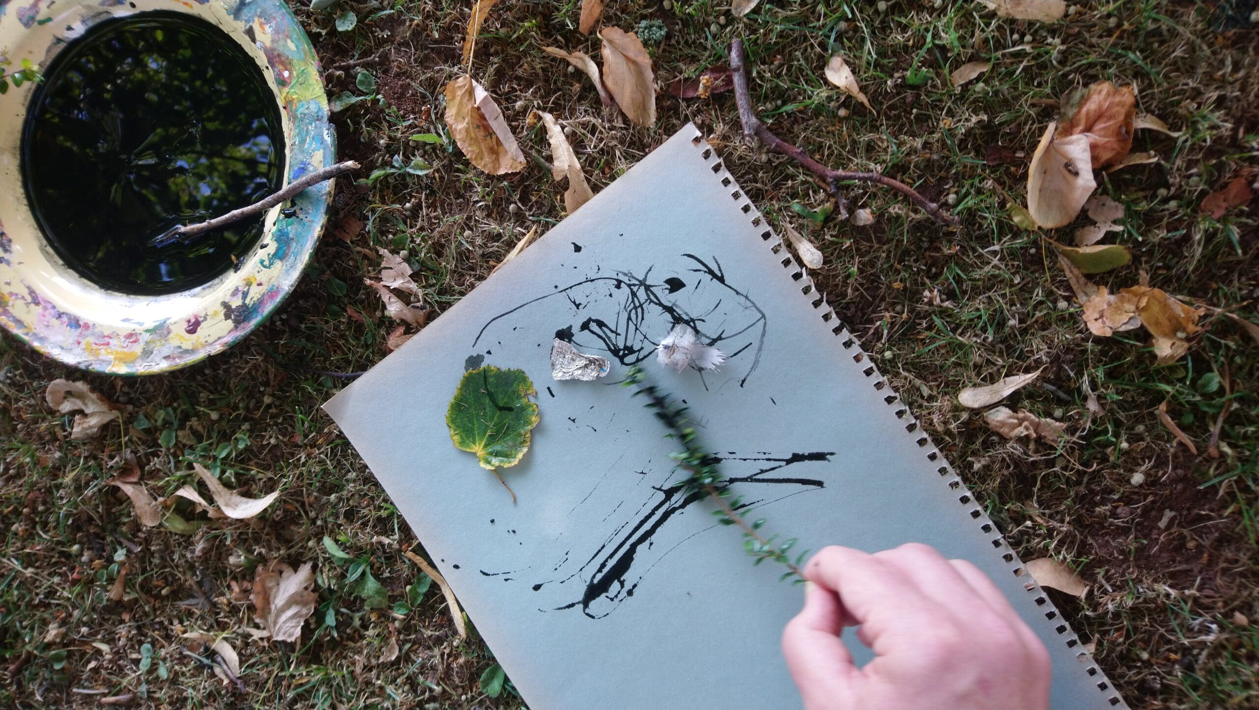 A hand uses a plant stem to paint black ink strokes on a sketchpad outdoors, with a bowl of ink, a leaf, and other natural objects nearby on the grass.