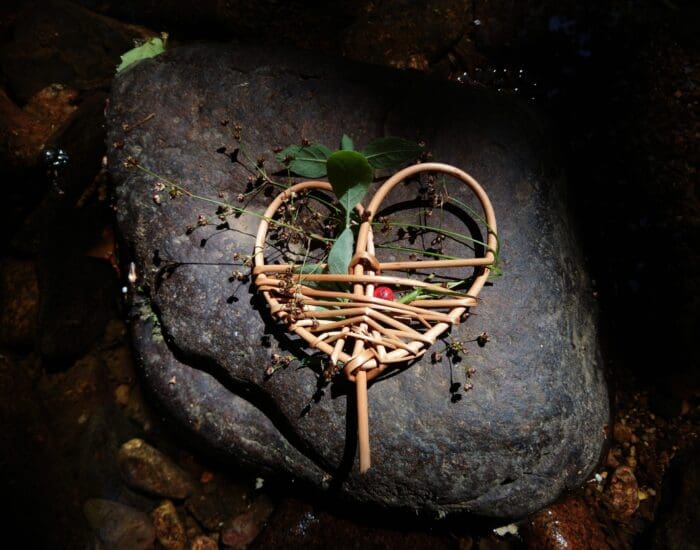 A woven heart-shaped ornament with twigs, leaves, and a red bead rests on a dark rock, surrounded by small plants and pebbles.