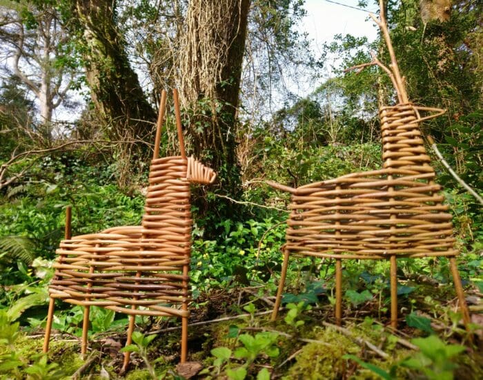 Two woven wicker animal sculptures, resembling a unicorn and a horse, stand on green foliage in a wooded outdoor setting.