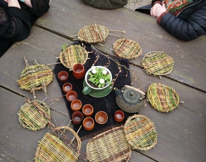 Three people sit around a wooden table set with woven placemats, a teapot, small cups, and a jug of water with floating flowers and leaves.