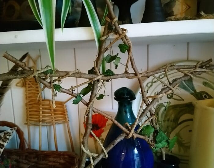 A handmade star-shaped frame of twigs and vines is displayed on a shelf with a blue glass bottle, three small black cups, pottery, and hanging green plant leaves.
