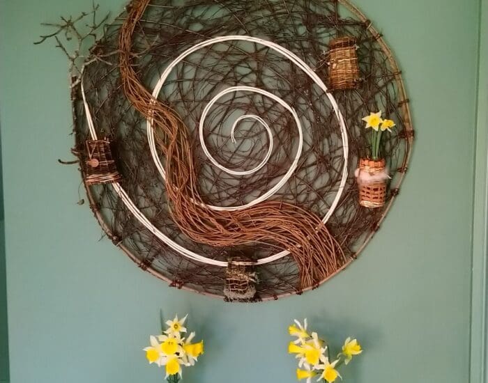 A round woven wall decoration with small baskets holding yellow flowers is mounted on a green wall above a wooden table with two small vases of yellow flowers.