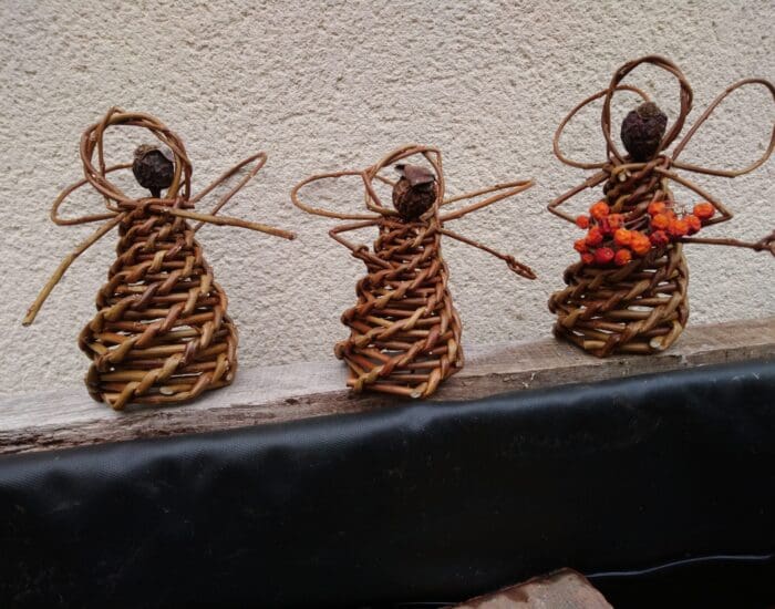 Three woven twig figures resembling angels are displayed on a wooden ledge against a textured wall; one holds a small bundle of orange berries.