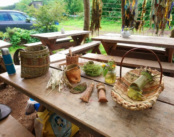 A wooden picnic table displays woven baskets, handmade crafts, a flask, flowers in jars, and weaving materials; benches and greenery are in the background.