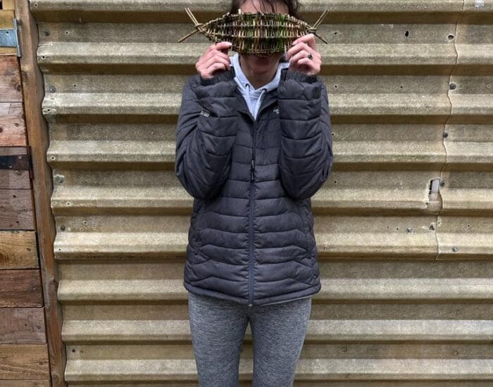 A person in a grey jacket and leggings stands against a corrugated metal wall, holding a stick with leaves in front of their face.