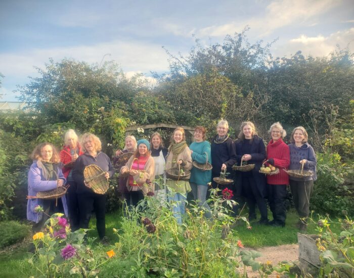A group of people stand outdoors in a lush garden, each holding a basket filled with harvested produce and flowers, smiling for the photo.
