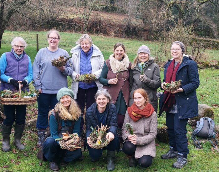 A group of ten people pose outdoors on grass, holding handmade woven baskets filled with natural materials, with trees and shrubs in the background.