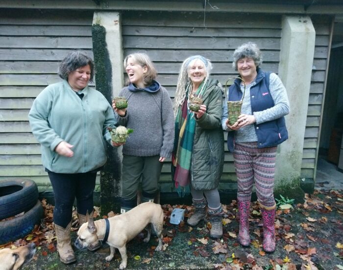Four women stand outdoors in front of a wooden wall, holding small potted plants. A dog stands near their feet on fallen leaves.