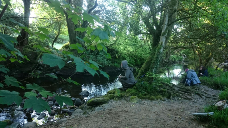 Several people sit and kneel by a forest stream, surrounded by trees and greenery, in a peaceful outdoor setting.